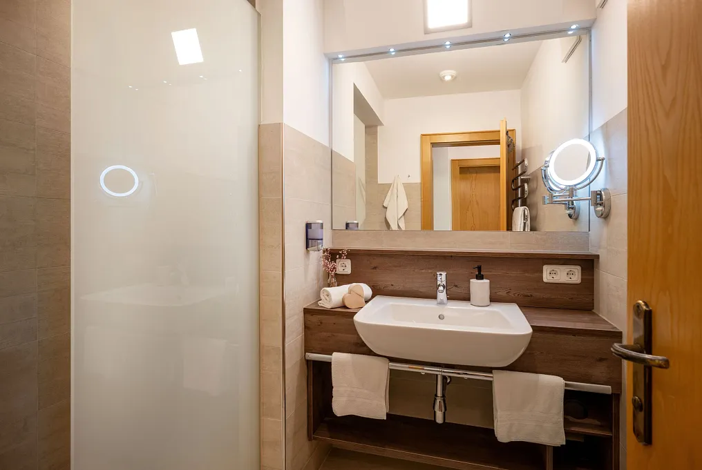 Modernes Badezimmer mit Waschbecken, beleuchtetem Spiegel und Handtüchern auf einem Holzregal.