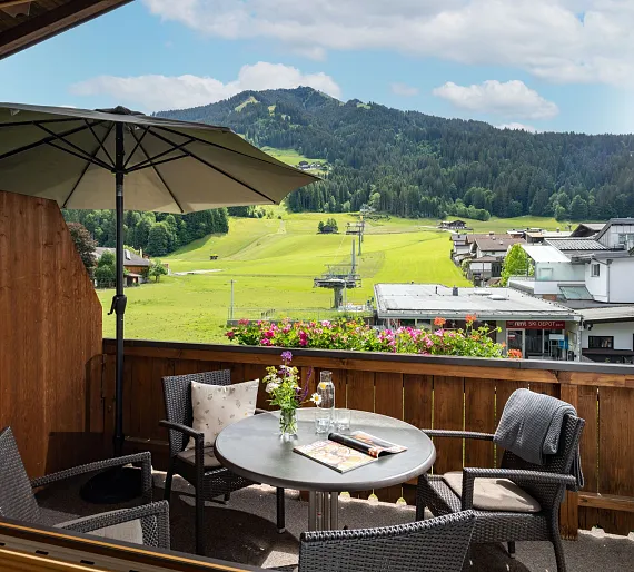 Balkon mit Tisch und Stühlen, Blick auf grüne Hügel und Seilbahn in Kitzbühel, Tirol bei klarem Himmel.