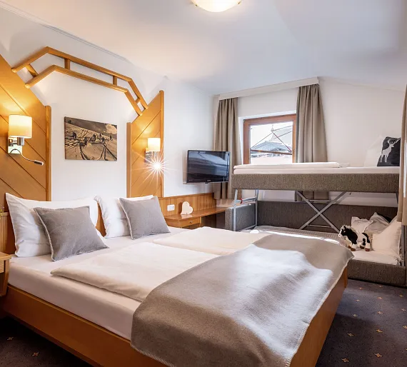 Hotelzimmer in Tirol mit Doppelbett, Hochbett, Fernseher und Fenster mit Vorhängen.