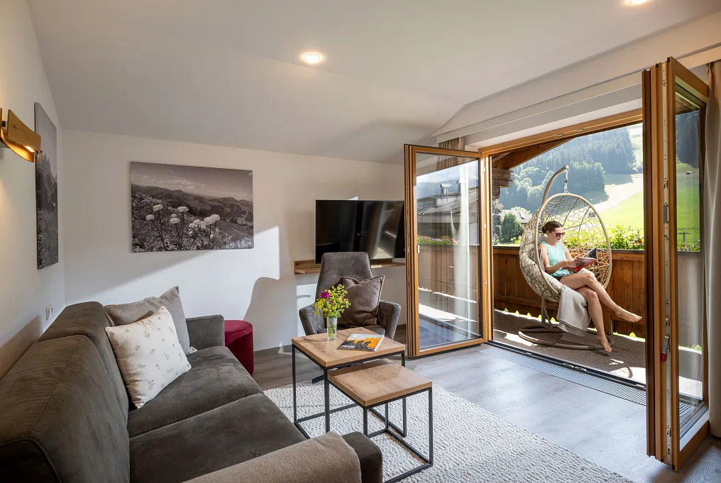 Wohnzimmer mit offener Balkontür, Frau in Hängesessel lesend, in alpiner Landschaft, moderne Ausstattung.