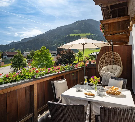 Balkon eines Hauses in Kitzbühel mit Blick auf die Berge und Blumen auf einem Geländer, bei sonnigem Wetter.