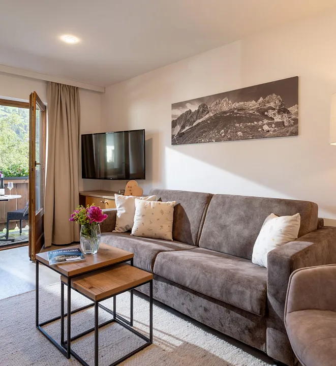 Wohnzimmer mit grauem Sofa, Couchtisch und TV, offen zu einem Balkon mit Blick ins Grüne.
