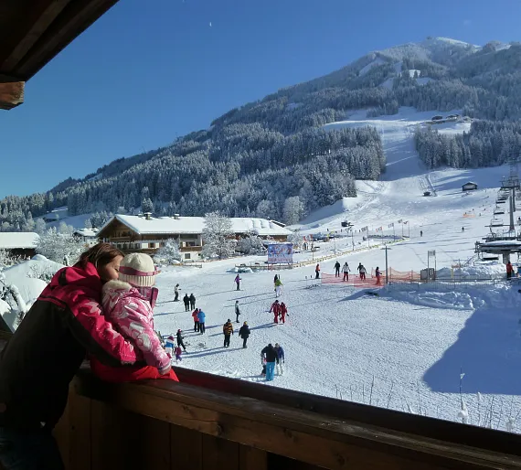Person hält ein Kleinkind auf einem Balkon mit Blick auf eine verschneite Skipiste in Tirol, Kitzbühel.