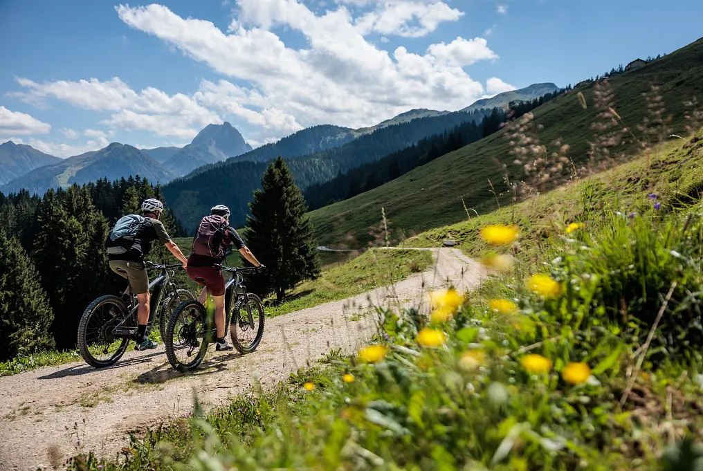 Zwei Radfahrer auf einem Bergweg vor alpiner Kulisse mit blühenden Wiesen in Tirol im Sommer.