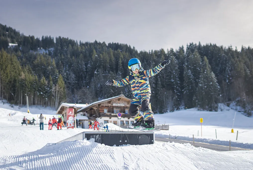 Kind auf einem Snowboard macht einen Trick vor einer Skihütte in verschneiter Berglandschaft.