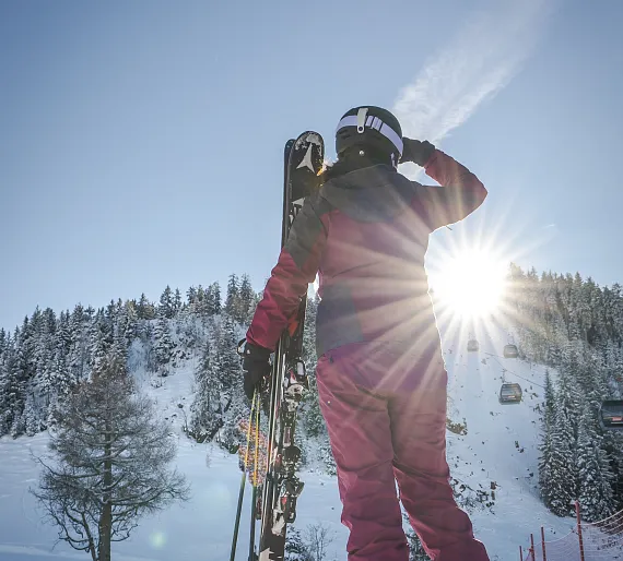 Person in Skikleidung hält Ski, blickt auf verschneite Berglandschaft mit Gondeln bei Sonnenschein.