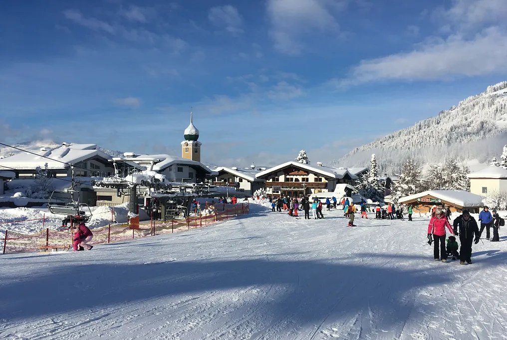 Verschneite Tiroler Berglandschaft mit Skifahrern vor einem alpinen Dorf mit Kirchturm.