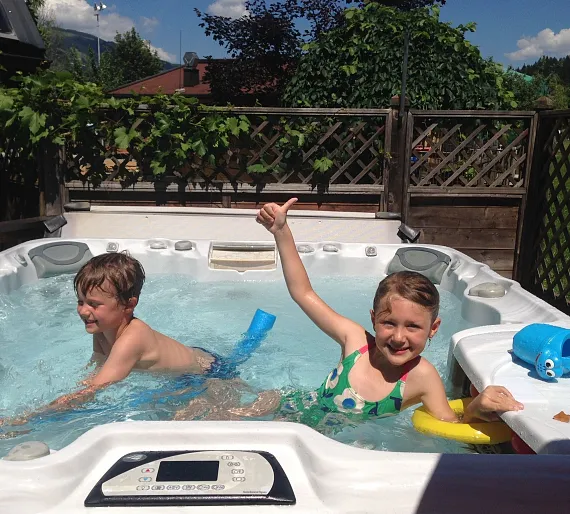 Zwei Kinder spielen in einem Whirlpool im Freien, umgeben von Holzzäunen und grüner Vegetation.