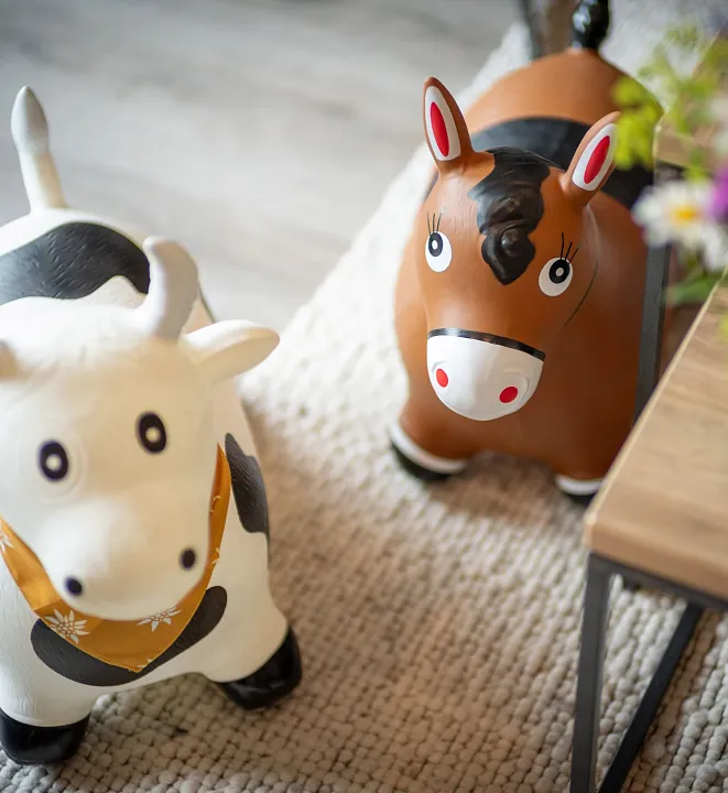 Zwei Spielzeug-Tiere, ein schwarz-weißer Bulle und ein braunes Pferd, stehen auf einem Teppich neben einem Holztisch mit Blumen.