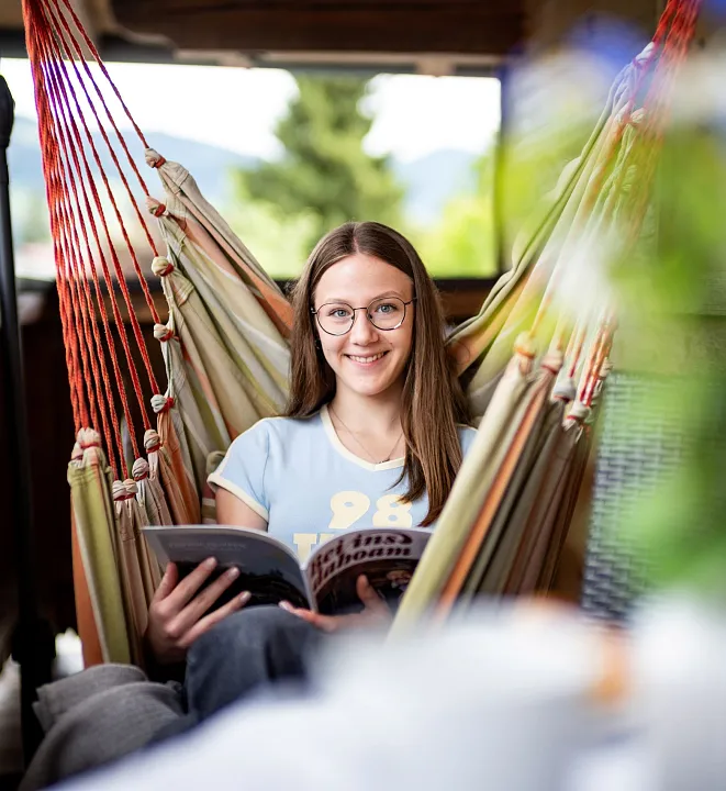 Junge Frau entspannt in einer Hängematte auf einem Balkon, liest ein Magazin; Berglandschaft im Hintergrund sichtbar.