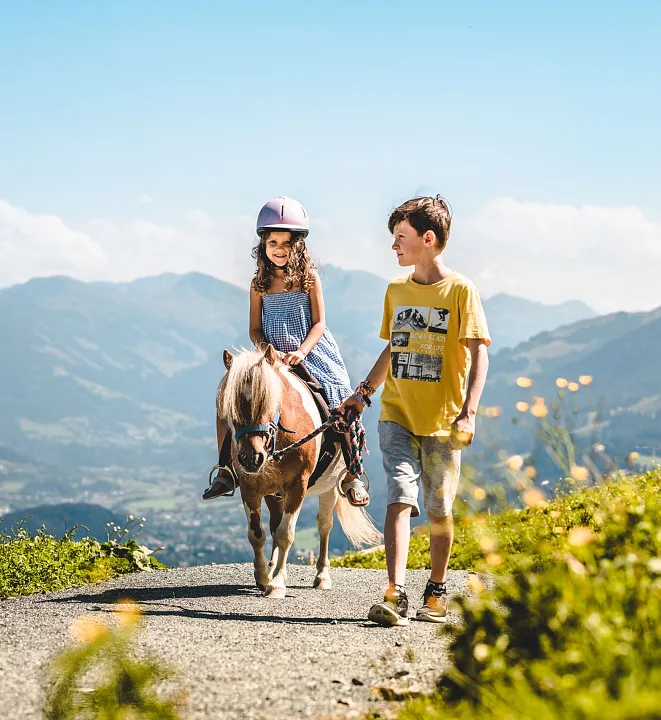 Mädchen auf Pony reitet in bergiger Landschaft, wird von einem Jungen geführt.