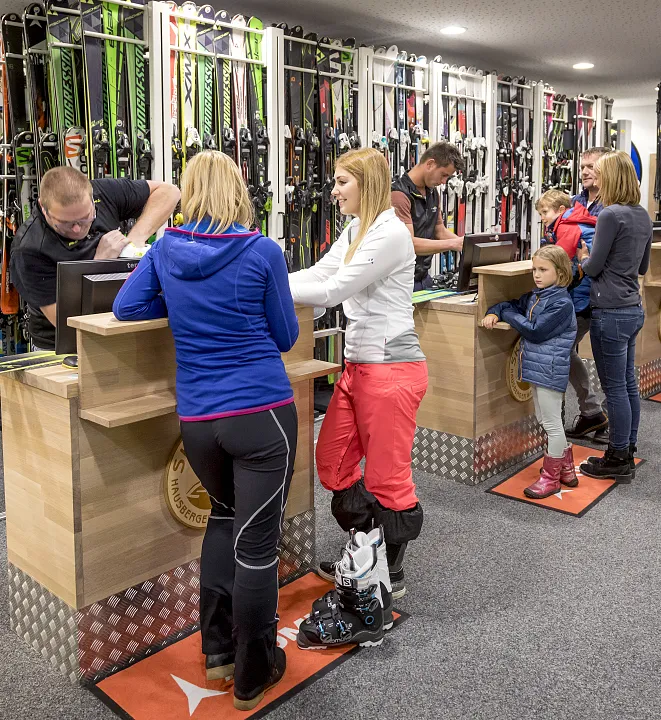 Menschen an einem Skiverleih-Tresen in einem Geschäft, zwei Frauen im Vordergrund leihen Skiausrüstung aus.