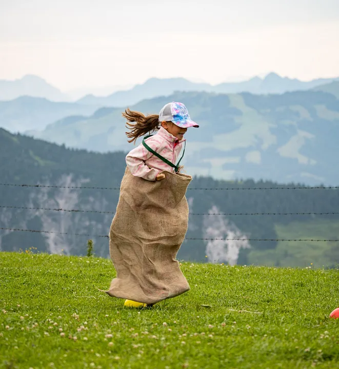 Kind mit Basecap hüpft in einem Sack auf einer Wiese in den Bergen.