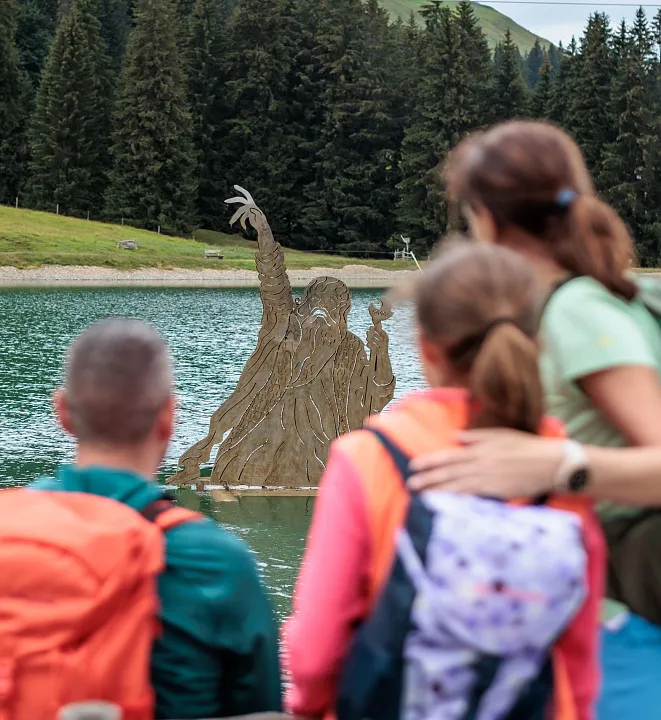 Drei Wanderer betrachten eine Skulptur in einem See in einer bewaldeten Berglandschaft.