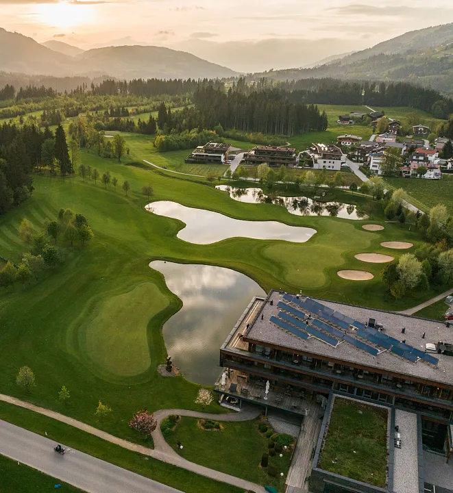 Golfplatz in einer bergigen Landschaft bei Sonnenuntergang, umgeben von Gebäuden und grünen Wiesen.