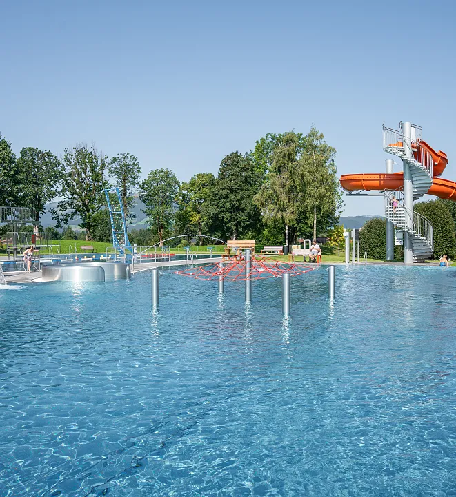Ein Freibad mit Wasserrutsche, mehreren Becken und Menschen an einem sonnigen Tag in einer grünen Umgebung.