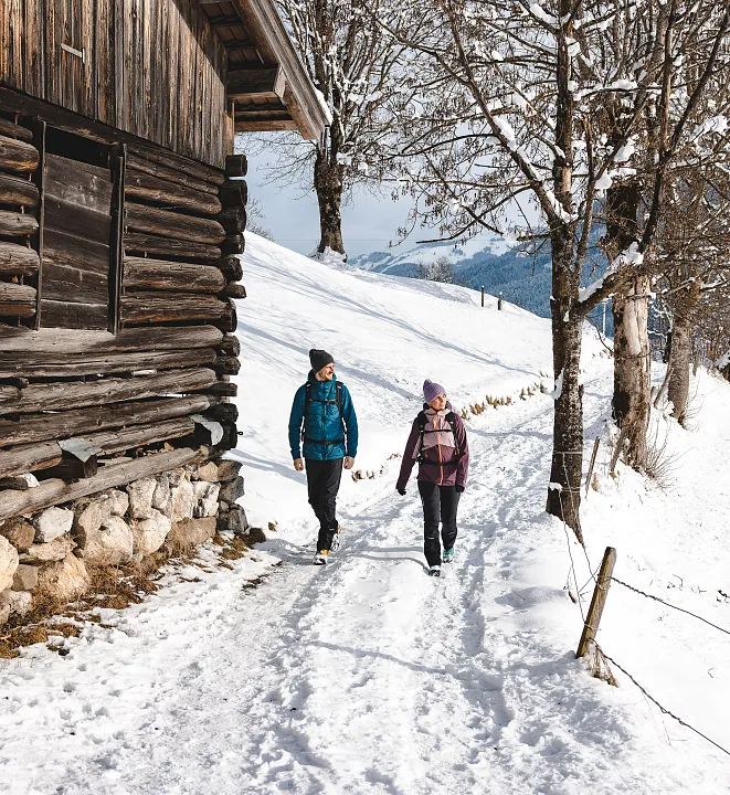 Zwei Personen wandern im Winter entlang eines schneebedeckten Pfades neben einer Holzhütte in Tirol, Kitzbühel.