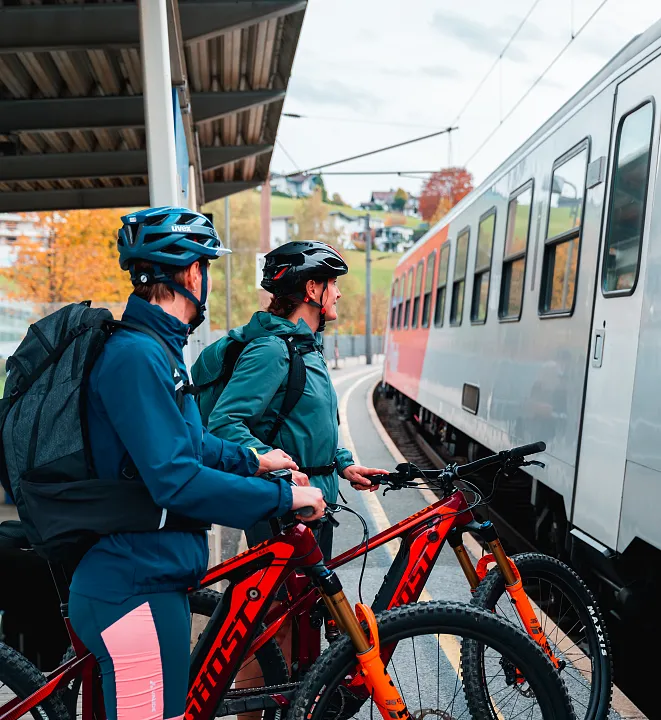 Zwei Personen mit Fahrrädern stehen auf einem Bahnsteig neben einem ÖBB-Zug in einer ländlichen Umgebung.