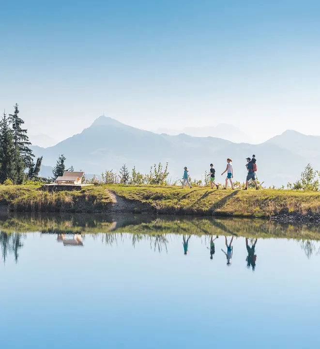 Personen spazieren entlang eines klaren Sees mit Berglandschaft im Hintergrund in einer alpinen Region.