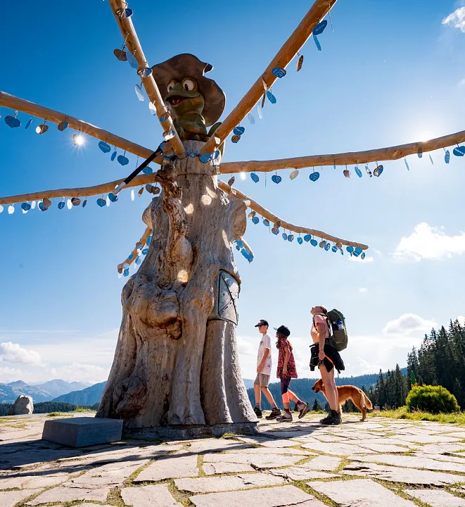 Drei Menschen wandern bei sonnigem Wetter an einem kunstvoll dekorierten Baum vorbei, im Hintergrund sind Berge und Wald zu sehen.