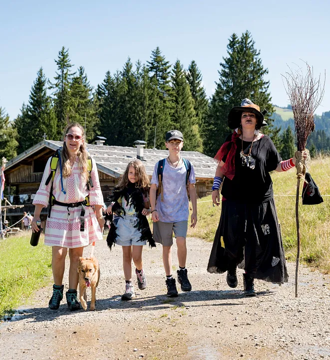 Vier Menschen und ein Hund spazieren auf einem Wanderweg in einer bergigen Waldlandschaft.