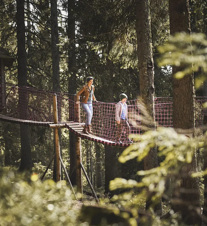 Zwei Personen gehen auf einer Hängebrücke in einem bewaldeten Gebiet.