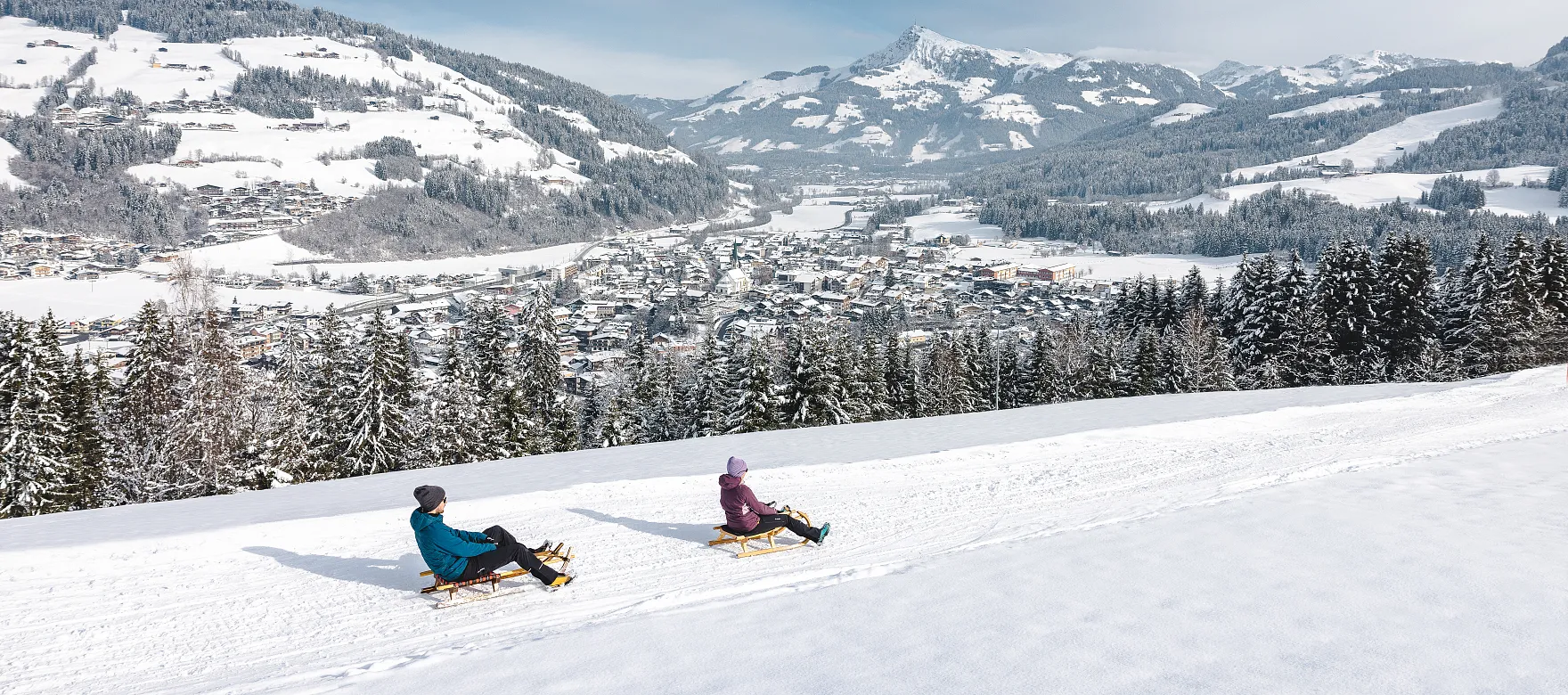 Zwei Personen rodeln auf einem verschneiten Hügel mit Blick auf die Berge und das winterliche Kitzbühel-Tal.