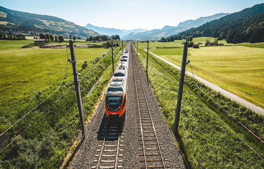 Regionalzug in Tiroler Landschaft auf gerader Strecke, umgeben von grünen Feldern und Bergen in der Ferne.
