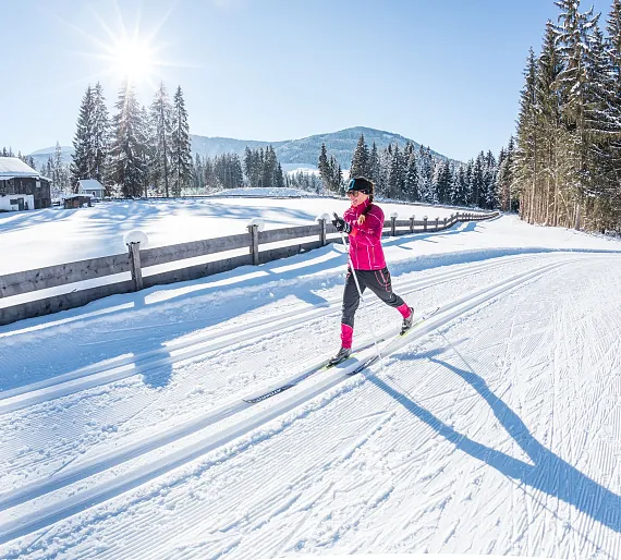 Langläuferin auf einer verschneiten Strecke in Tirol bei Sonnenschein, umgeben von Wäldern und Bergen.