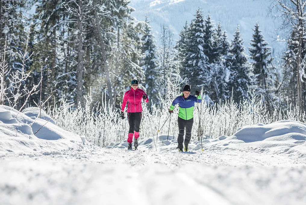 Zwei Personen beim Wintersport auf einer verschneiten Langlaufloipe in einer Waldlandschaft.