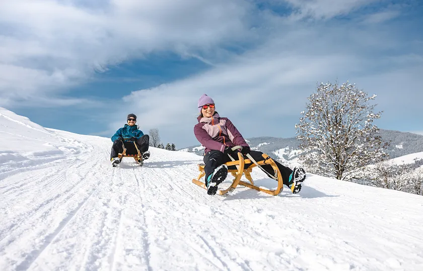 Zwei Personen rodeln auf einem schneebedeckten Hang an einem sonnigen Tag.
