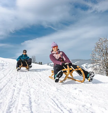 Zwei Personen rodeln auf einem schneebedeckten Hang an einem sonnigen Tag.