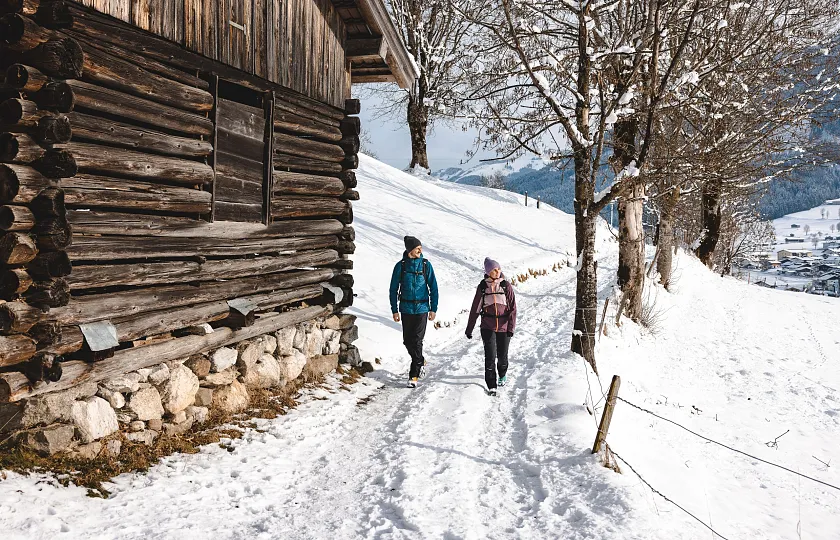Zwei Personen wandern im Winter entlang eines schneebedeckten Pfades neben einer Holzhütte in Tirol, Kitzbühel.
