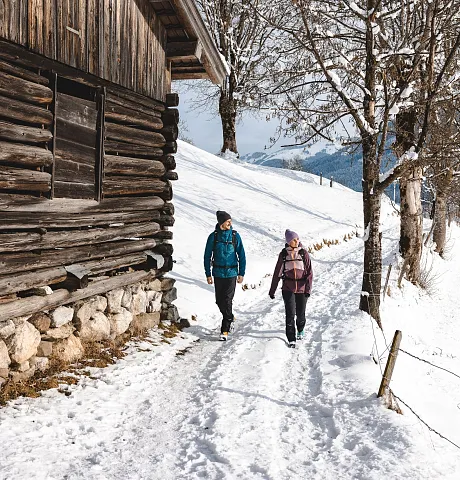 Zwei Personen wandern im Winter entlang eines schneebedeckten Pfades neben einer Holzhütte in Tirol, Kitzbühel.