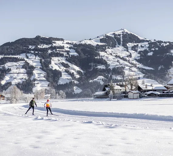 Zwei Langläufer in winterlicher Landschaft mit schneebedeckten Bergen im Hintergrund, nahe Kitzbühel, Tirol.