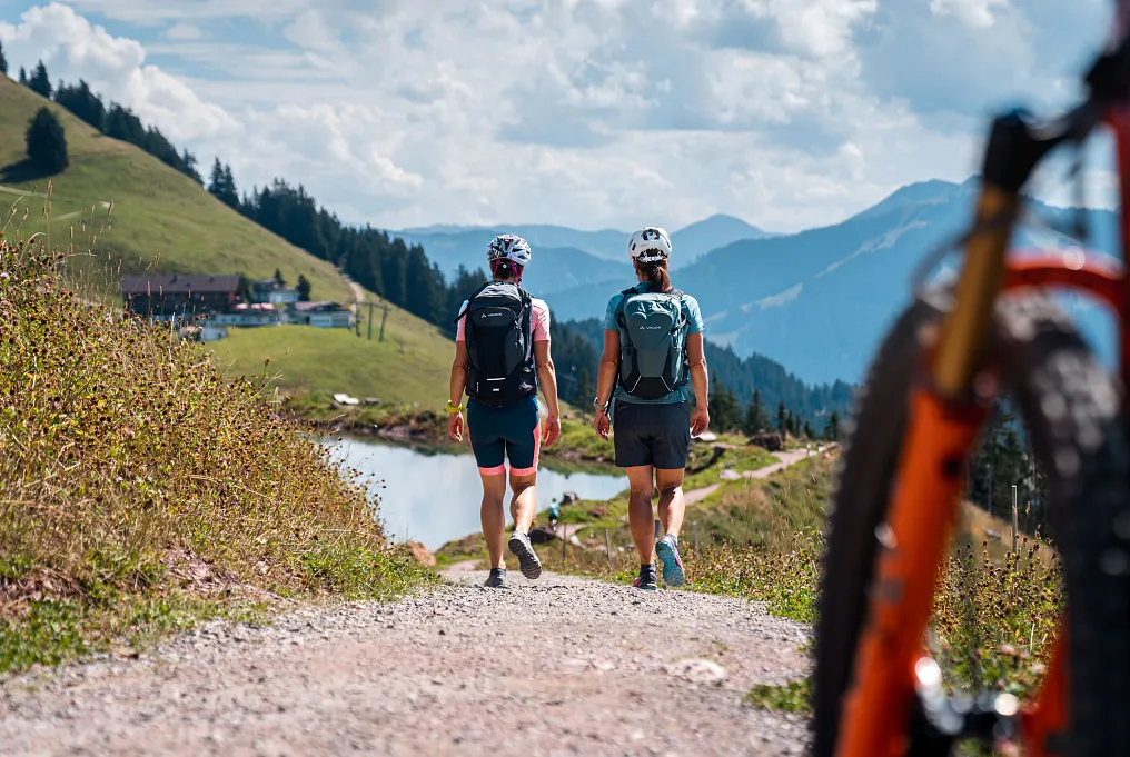 Zwei Personen beim Wandern in bergiger Landschaft mit Blick auf Tiroler Alpen, ein Fahrrad am Bildrand.