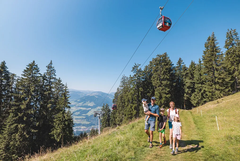 Familie spaziert auf einem Wanderweg in Tirol, eine Seilbahn fährt darüber hinweg, und im Hintergrund sind Berge sichtbar.