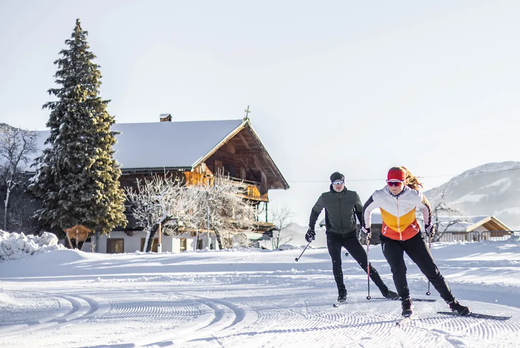 Zwei Langläufer gleiten auf einer verschneiten Loipe vor einem traditionellen Holzhaus in Tirol.
